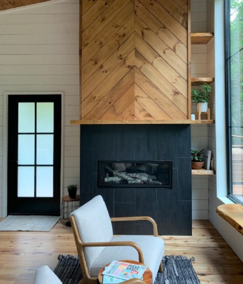 Cozy interior featuring a wooden fireplace, modern chair, and large windows with greenery outside.