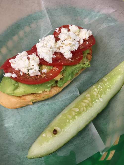 A sandwich topped with avocado, tomatoes, and feta cheese, served with a slice of cucumber on the side.