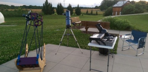 A grassy area with telescopes set up on a patio, surrounded by trees and a distant building under a cloudy sky.