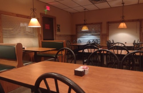 Empty restaurant interior with wooden tables, green benches, and warm lighting. Windows covered with blinds.
