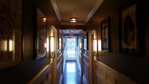 A dimly lit hallway with framed pictures and wall sconces, leading to a door at the end.