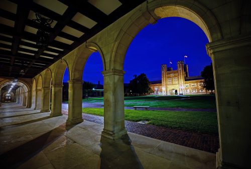 A nighttime view of a university campus, featuring arched stone walkways and a lit building in the background.