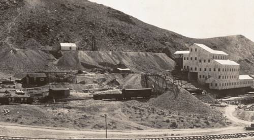 Historic mining site with buildings, equipment, and rail cars, set against a mountainous landscape.