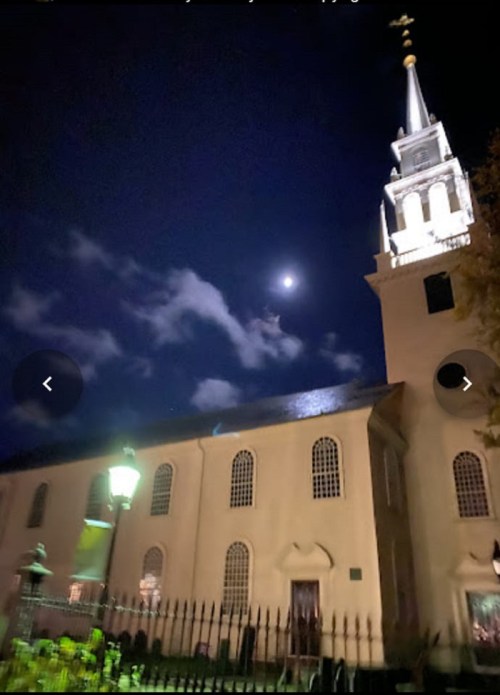 A church at night with a lit steeple and a bright moon in the sky, surrounded by clouds and a lamppost.