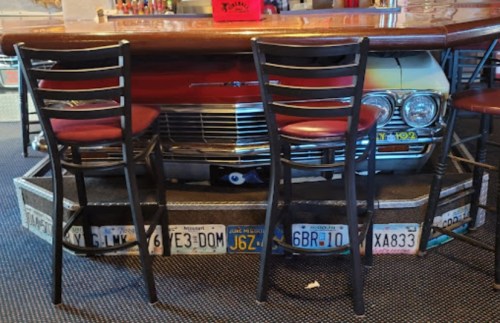 A vintage car is partially visible under a bar counter, surrounded by bar stools and decorated with license plates.