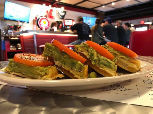 A plate of avocado toast topped with tomato slices, served in a diner with a lively background.