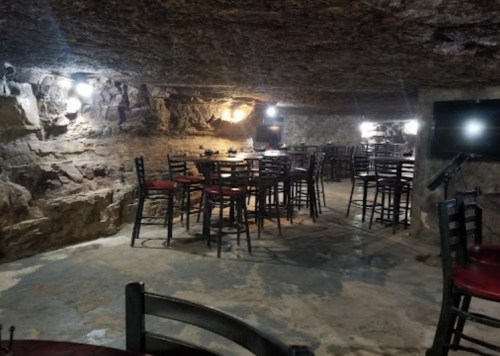 A dimly lit underground space with stone walls, featuring tables and chairs arranged for dining.