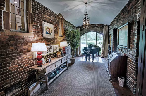 A cozy hallway with brick walls, a piano, lamps, and decorative plants, leading to a bright arched window.