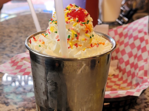 A metal cup filled with whipped cream, colorful sprinkles, and a cherry on top, with two straws.