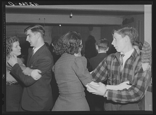 A group of people dancing in a dimly lit room, dressed in formal and casual attire, enjoying a social event.