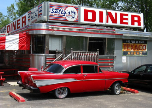 A classic red car parked outside a retro diner with a shiny silver exterior and a bright sign.