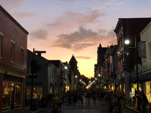 A bustling street at sunset, lined with shops and crowds, under a colorful sky with clouds.