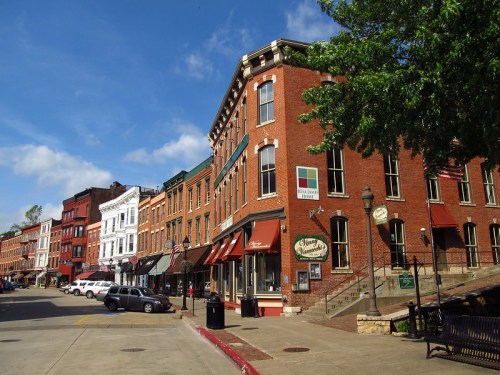 Historic brick buildings line a street, featuring shops and cafes under a clear blue sky.