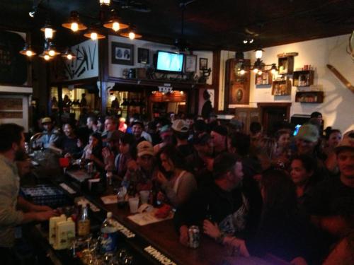 A crowded bar scene with patrons socializing, drinks on the counter, and a bartender serving in a lively atmosphere.