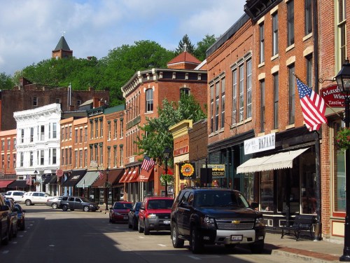 A charming street lined with historic brick buildings, shops, and American flags, under a blue sky with greenery above.