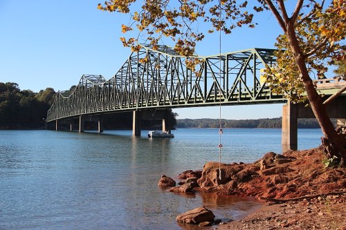 A green metal bridge spans a calm lake, with rocky shores and trees in the foreground under a clear blue sky.