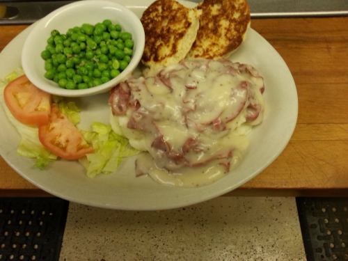 A plate of mashed potatoes topped with creamy meat, served with green peas, lettuce, tomato slices, and toasted bread.