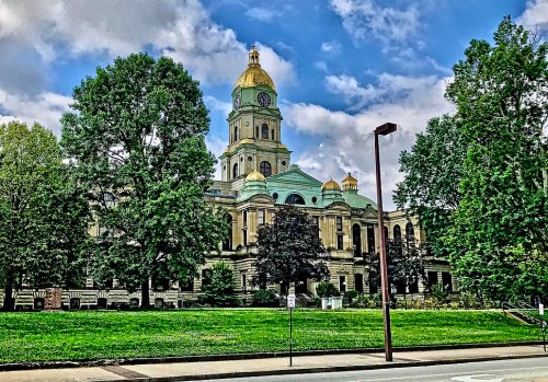 Historic building with a golden dome, surrounded by trees and a grassy area under a blue sky with clouds.