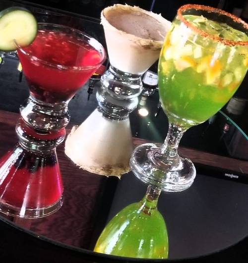 Three colorful cocktails on a reflective surface: a red drink with a cucumber slice, a creamy white drink, and a green drink.