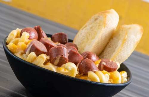 A black bowl filled with macaroni and cheese topped with sliced hot dogs, accompanied by two pieces of bread.