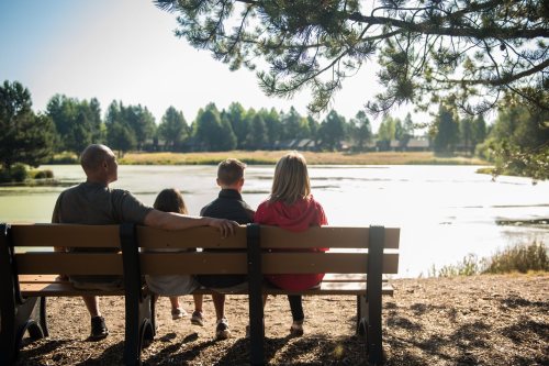 A family of four sits on a bench by a serene lake, enjoying the view of trees and water on a sunny day.