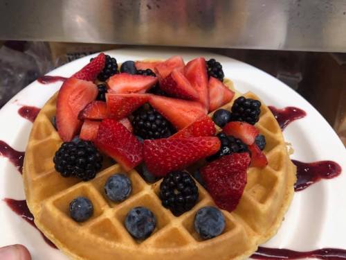 A golden waffle topped with fresh strawberries, blackberries, and blueberries, drizzled with berry sauce.