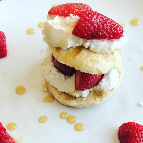 A layered dessert with strawberries, whipped cream, and a biscuit, drizzled with honey on a white plate.