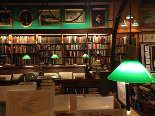A cozy library filled with bookshelves, green lamps, and framed artwork on the walls, creating a warm reading atmosphere.
