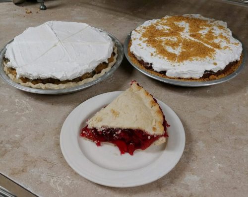 Three pies on a countertop: two whole pies with whipped topping and a slice of cherry pie on a plate.