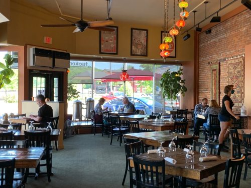 A cozy restaurant interior with wooden tables, plants, and patrons dining by a large window.