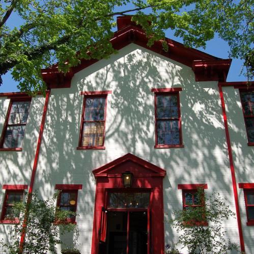 A white building with red trim and windows, surrounded by green trees casting shadows on the facade.