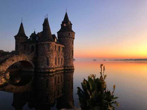 A stone castle with towers reflects in calm water at sunrise, surrounded by a serene landscape.