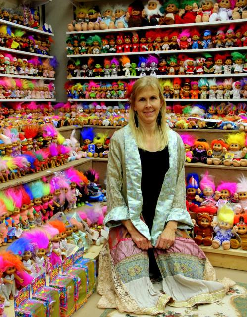 A woman sits among colorful troll dolls on shelves, wearing a long, patterned dress and a light shawl.
