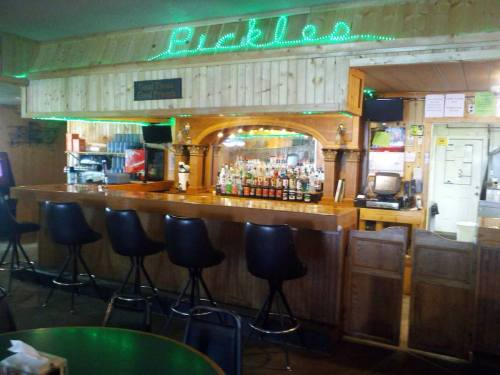 A rustic bar with a wooden counter, high stools, and a well-stocked shelf of liquor bottles, illuminated by green lights.