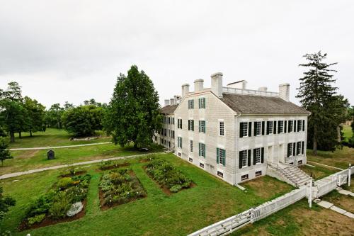 Historic white house with green shutters, surrounded by gardens and trees, under a cloudy sky.