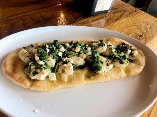 A flatbread topped with shrimp, greens, and crumbled feta cheese, served on a white plate.
