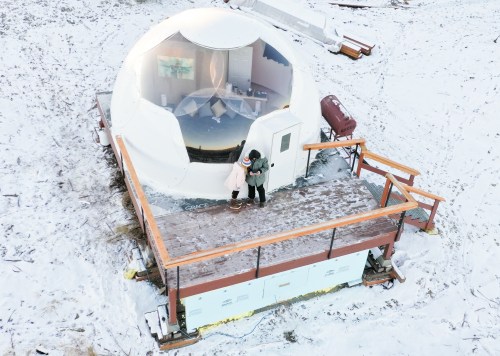 A couple stands on a snowy deck in front of a geodesic dome, surrounded by winter scenery.