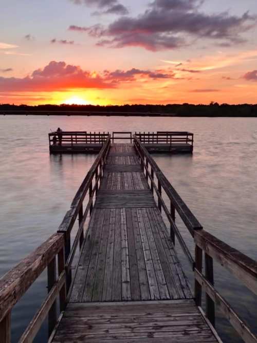 A wooden pier extends into a calm lake at sunset, with vibrant colors reflecting on the water.