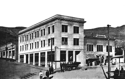 Historic black-and-white photo of a multi-story building at a street corner, with early 20th-century vehicles and pedestrians.