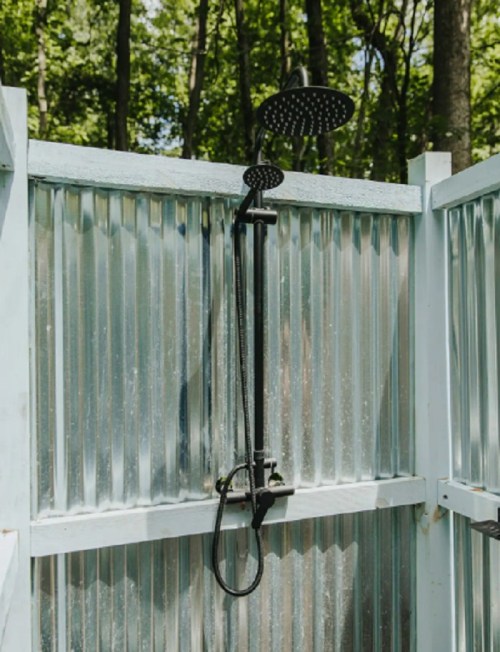 Outdoor shower with a black showerhead, surrounded by corrugated metal walls and greenery in the background.