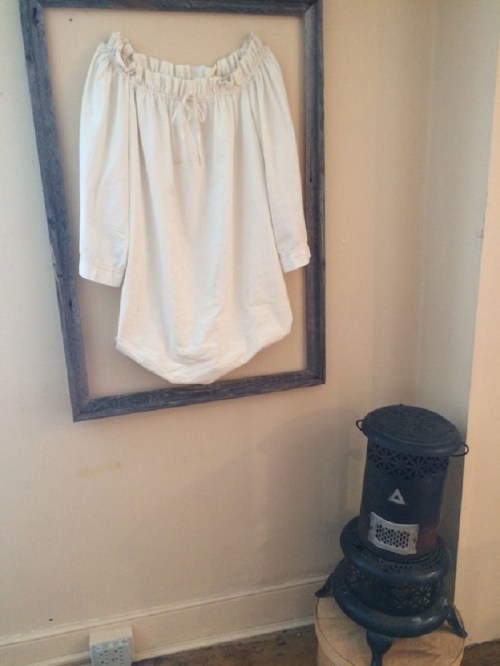 A white, vintage-style shirt displayed in a wooden frame next to a black, decorative stove.