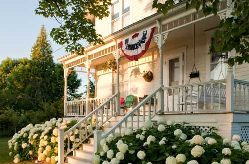 A charming white house with a porch, decorated with bunting, surrounded by blooming hydrangeas in the foreground.