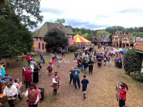 A lively outdoor festival scene with people in costumes, colorful tents, and medieval-style buildings.