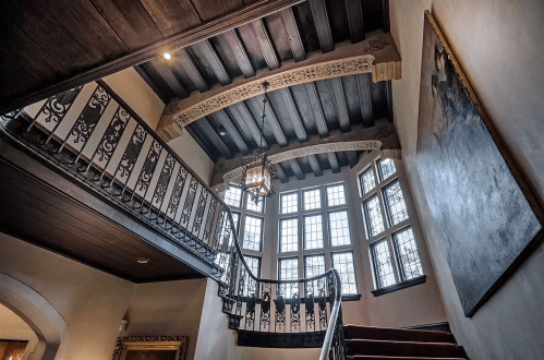 Elegant staircase with ornate railing, wooden beams, and large windows, illuminated by a vintage lantern.