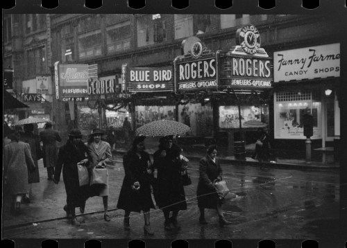 Black and white photo of people walking in the rain, passing storefronts with neon signs and umbrellas.