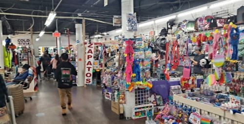 A bustling market aisle filled with colorful merchandise, including toys, accessories, and clothing, with shoppers browsing.