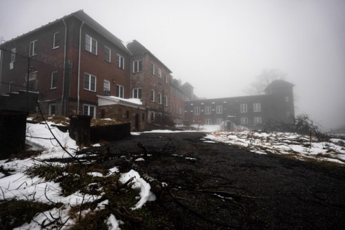 Abandoned building shrouded in fog, with snow and overgrown vegetation in the foreground.