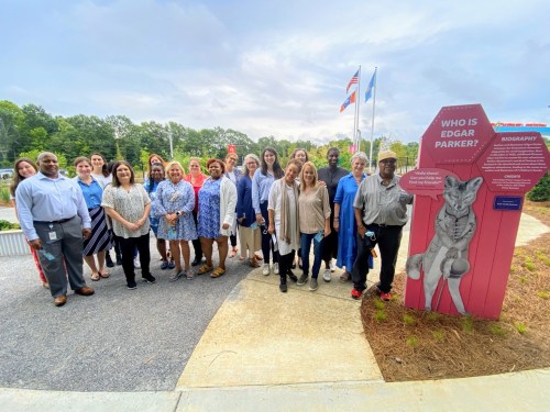A diverse group of people poses outdoors near a sign about Edgar Parker, with flags and trees in the background.