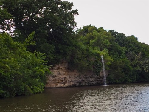 A serene river scene featuring a waterfall cascading from a lush, green cliff into the calm water below.