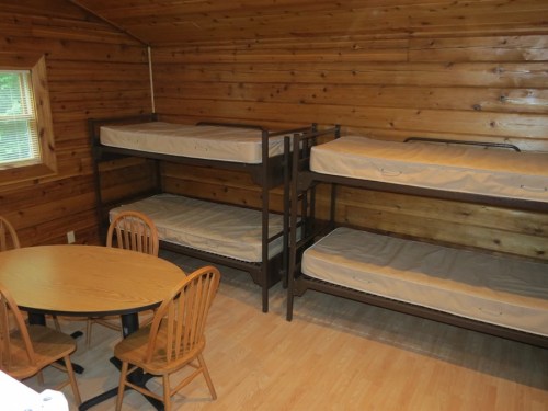 A rustic cabin interior featuring two sets of bunk beds and a round wooden table with four chairs.